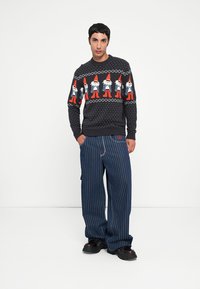 Schwarzer gestrickter Pullover mit festlichem Gnome-Design, kombiniert mit einer lockeren blauen Nadelstreifenhose und dunklen Schuhen. Weiße Punkte auf dem Pullover.