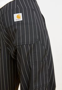 Mustat raidoitetut housut, jotka on valmistettu sileästä kankaasta. Varustettu taskulla ja näyttävällä Carhartt-logo-patchilla takana.