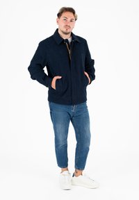 Giacca in tessuto blu navy con taglio dritto, dotata di due tasche frontali e colletto. Abbinata a jeans azzurri chiari e sneakers bianche.