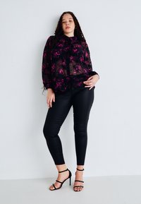 Blouse noire transparente avec imprimé floral rose, manches longues et fermeture à boutons, associée à un pantalon skinny noir et des escarpins noirs à lanières.