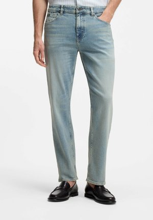 Mann trägt hellblaue Denim-Jeans und schwarze Leder-Loafers, steht mit einer Hand in der Tasche vor weißem Hintergrund.