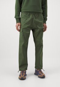 Gröna cargobyxor med elastisk midja, sidofickor och avsmalnande ben, tillverkade av lätt tyg, som bärs med bruna och blå skor.