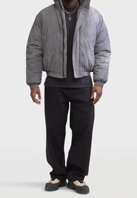 Grå quiltet bomberjakke med ribbede manchetter, båret over en sort t-shirt og løse sorte bukser. Kraftige beige sneakers fuldender outfittet.