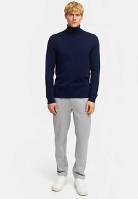 Granatowy golf wykonany z lekkiego dzianinowego materiału, zestawiony z jasnoszarymi spodniami dresowymi. Model ma na sobie białe trampki, stoi na jednolitym tle.