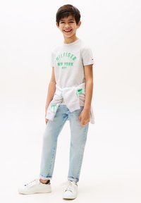Camiseta gris con "HILFIGER NEW YORK CITY" en verde, jeans claros, y zapatillas blancas. Suéter atado a la cintura. Fondo minimalista.