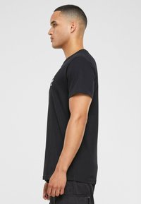 Sort bomulds T-shirt med korte ærmer, rund hals og logodesign, præsenteret iført af en person i profil mod en lys baggrund.