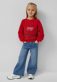 Jong meisje staat met handen in haar zakken, draagt een rode sweatshirt met een zwaan grafiek en tekst, wijde blauwe jeans en witte sneakers.