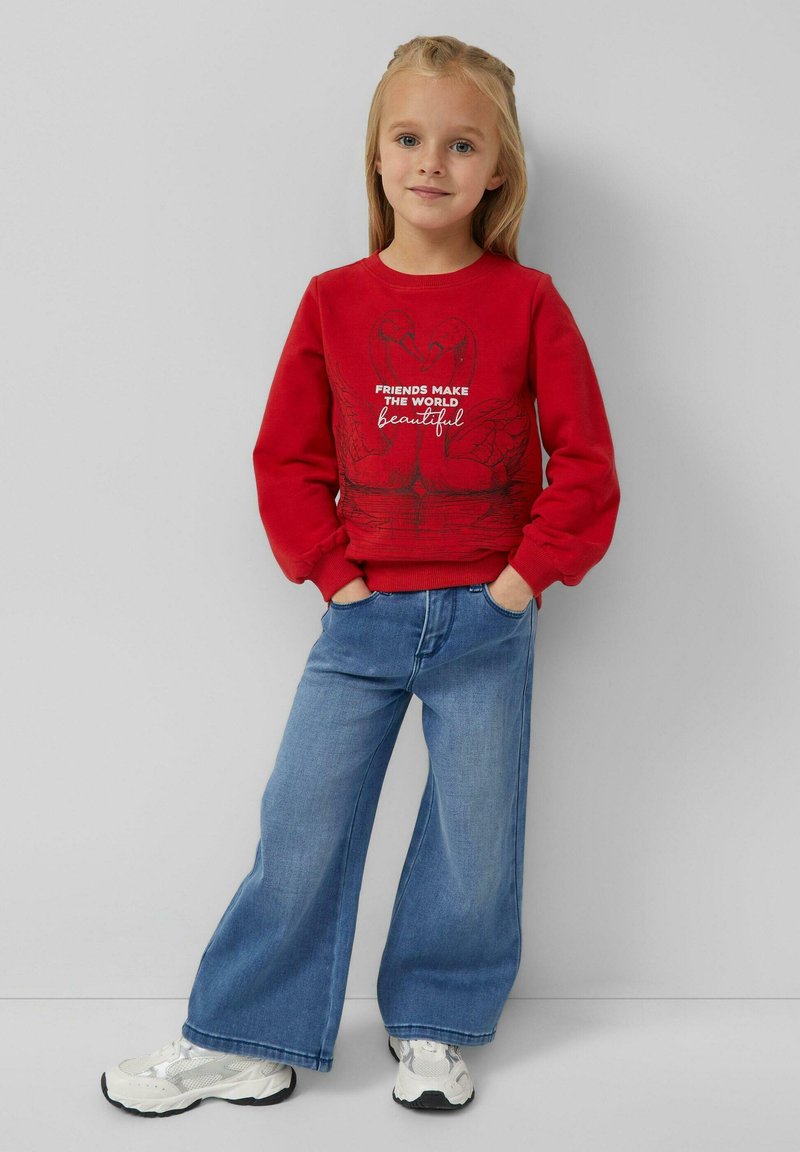 Jong meisje staat met handen in haar zakken, draagt een rode sweatshirt met een zwaan grafiek en tekst, wijde blauwe jeans en witte sneakers.