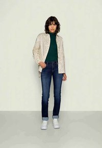 Chemise boutonnée beige à motifs sur un pull à col roulé vert, associée à un jean bleu foncé et des baskets blanches, se tenant sur un fond neutre.