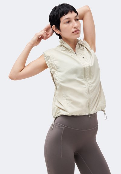 Άτομο με κοντά σκούρα μαλλιά που τεντώνει τα χέρια πίσω από το κεφάλι, φοράει αμάνικο ανοιχτό γιλέκο με φερμουάρ και γκρι κολάν με ψηλή μέση.