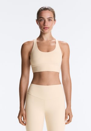 Vrouw met haar naar achter gebonden, gekleed in een beige sportbeha en hooggetailleerde legging, staand tegen een effen lichte achtergrond, naar voren kijkend.