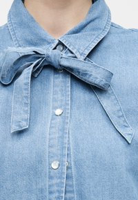 Γαλάζιο τζιν πουκάμισο με μπροστινή σειρά κουμπιών και υφασμάτινο φιόγκο δεμένο στο γιακά.