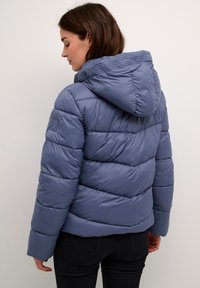 Lightweight puffer jacket in muted blue, featuring a hood, horizontal stitching, and a snug fit. Material appears smooth and glossy.