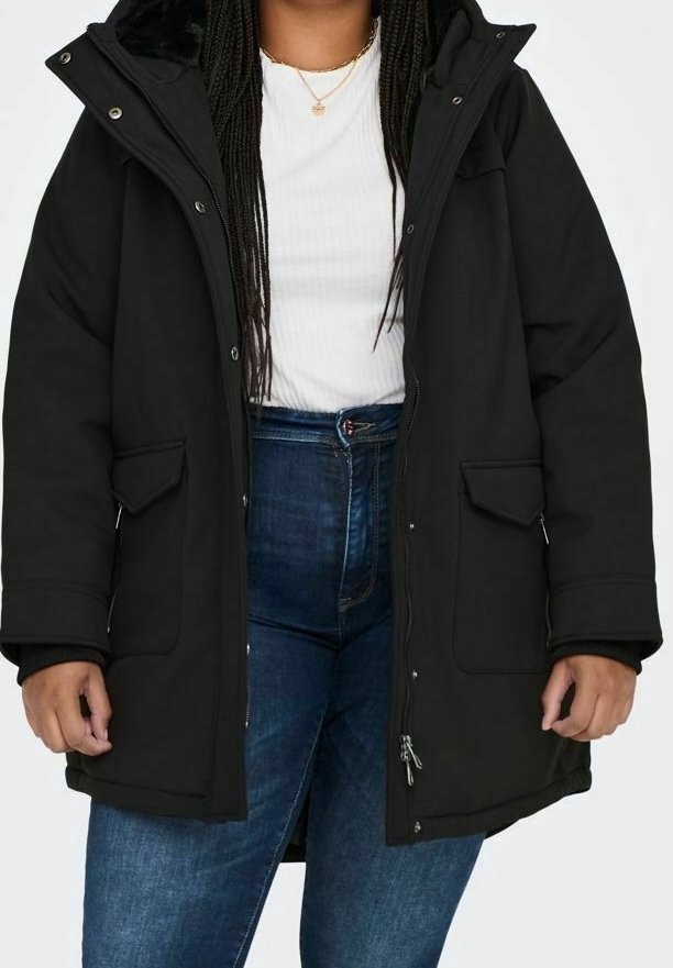 Personne portant une veste d'hiver noire à capuche sur un haut blanc et un jean bleu taille haute, debout les mains détendues le long du corps.
