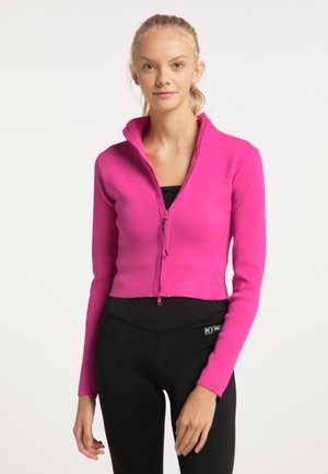 Junge Frau trägt eine leuchtend pinke, kurze Jacke mit Reißverschluss über einem schwarzen Top und schwarzen engen Hosen und steht vor einem schlichten weißen Hintergrund.