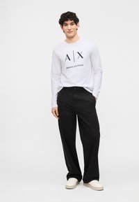 Άνδρας που στέκεται με τα χέρια στις τσέπες, φορώντας λευκό μακρυμάνικο πουκάμισο Armani Exchange, μαύρο παντελόνι και λευκά αθλητικά παπούτσια, με φόντο απλό.