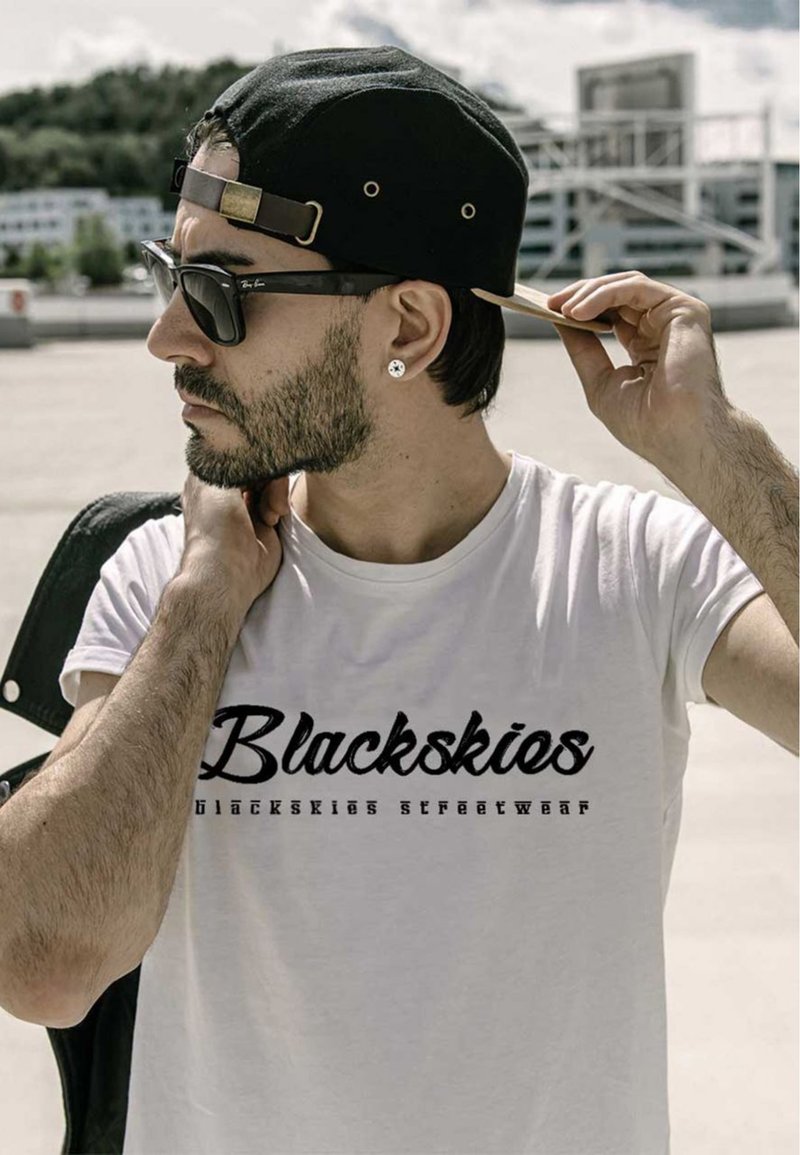 Weißes Baumwoll-T-Shirt mit einem schwarzen Grafikdruck, auf dem "Blackskies streetwear" steht. Das Model trägt Sonnenbrillen und eine schwarze Mütze.