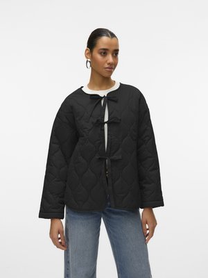 Femme portant une veste matelassée noire avec trois nœuds papillon, chemise blanche, boucles d'oreilles créoles et jean bleu clair, debout devant un fond uni.