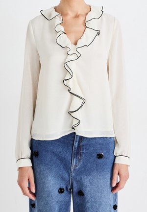 Weiße Bluse mit langen Ärmeln und schwarzem, mit Rüschen besetztem Saum auf der Vorderseite, kombiniert mit blauen Jeans, die schwarze Blumenverzierungen aufweisen.