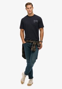 Mann trägt schwarzes T-Shirt mit Grafik, blaue Jeans mit karierter Hemd um die Taille gebunden, beige Sneakers und Armbanduhr, steht mit den Händen in den Taschen.