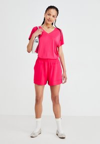 Roze, kortmouw V-hals top gecombineerd met roze shorts met voorkruipzakken. Model houdt een zilveren tas vast, draagt witte sokken en platte schoenen.