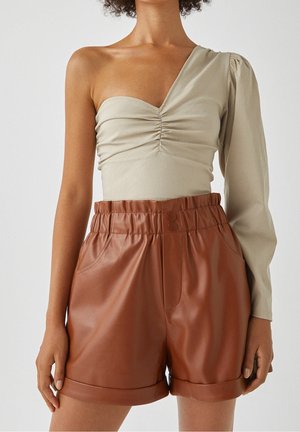 Femme portant un haut beige asymétrique à une épaule à manches longues et un short en cuir marron taille haute avec revers roulés et taille élastique.