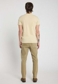 T-shirt beige à manches courtes et pantalon marron clair avec un détail de poche arrière. Mélange de coton, texture douce, porté avec des chaussures marron.