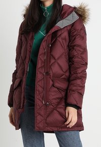 Bordeaux pudejakke med pelsfor i hætten, grå detaljer, to forlommer og lynlås/knap lukning. Båret over en grøn top og jeans.