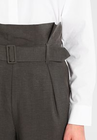 Spodnie z wysokim stanem w teksturowanej, ciemnoszarej tkaninie z szerokim paskiem i detalem klamry, zestawione z białą koszulą z guzikami.