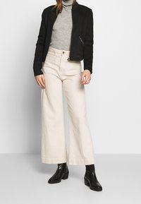 Giubbotto bomber in suede nero, dolcevita grigio a coste, pantaloni beige a gamba larga e stivaletti neri. Design semplice con dettagli hardware minimi.