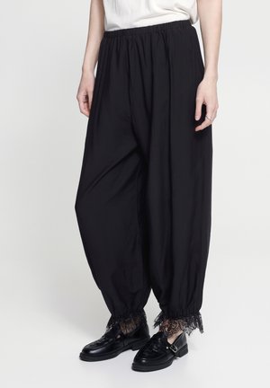 Široké čierne nohavice s elastickým pásom a čipkovaným lemom pri členkoch, nosené s čiernymi mokasínami a bielym topom na stojacej osobe.