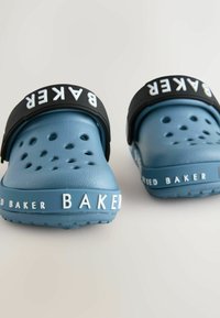 Modré gumené topánky s textúrovaným povrchom, vybavené ventilačnými otvormi. Čierny remienok so slovom "BAKED" v bielej farbe, ktoré je výrazne zobrazené.