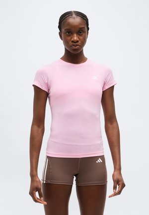 Nő, aki szűk, világos rózsaszín rövid ujjú sportfelsőt és barna rövidnadrágot visel fehér csíkokkal, egyszerű háttér előtt áll.