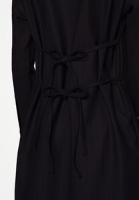 Svart tygkappa med vertikala randiga mönster, med knytfäste i midjan, långa ärmar och en slät yta.