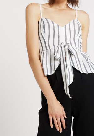 Vrouw draagt een wit en grijs verticaal gestreept mouwloos topje met een voorstrik, gecombineerd met zwarte broeken met hoge taille tegen een effen achtergrond.