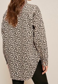 Camicia a fantasia leopardata con base beige chiaro e macchie nere, maniche lunghe e orlo curvo, che mostra una texture del tessuto liscia.