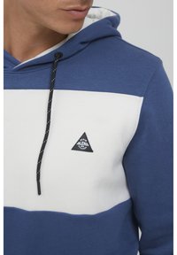 Felpa con cappuccio blu e bianca, caratterizzata da un logo triangolare, lacci neri con dettagli bianchi e una finitura in tessuto testurizzato.