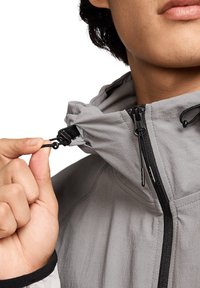 Grijze jas met een capuchon, voorzien van een verstelbare koord, zwarte ritsaccenten en een soepele textuur. Close-up van een hand die de kraag aanpast.