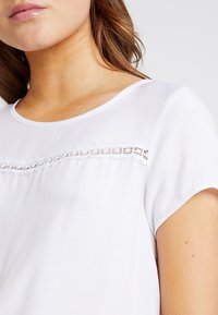 Weißes Kurzarmoberteil mit glatter Textur, rundem Halsausschnitt und dekorativem Spitzeinsatz über der Brust. Einfaches und klares Design.