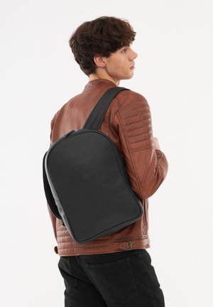 Mochila de cuero negro con forma redondeada, textura suave y cierre de cremallera, llevada en un hombro sobre una chaqueta de cuero marrón.