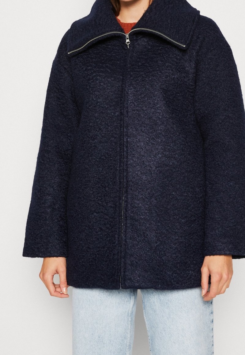 Tummansinisestä, teksturoituneesta kankaasta valmistettu ylipitkä takki, jossa on korkea kaulus ja etuvetoketju, yhdistettynä vaaleansinisiin farkkuihin.