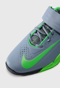 Grå atletiksko med en tekstureret overdel, grønne accenter, en Velcro-rem og perforerede detaljer for åndbarhed. Nike-logo i grøn.