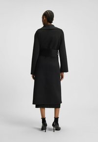 Black long coat with a collar and a belted waist, featuring long sleeves and a straight silhouette, worn over a black ribbed skirt and heels.