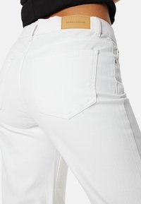 Jeans in denim bianco con una vista ravvicinata, dotati di cinque tasche, una patch in pelle del marchio sulla vita e un design aderente.