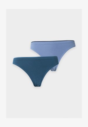 Femme debout pieds nus portant un soutien-gorge gris clair et une culotte bleue, une main sur la hanche, sur un fond blanc uni.