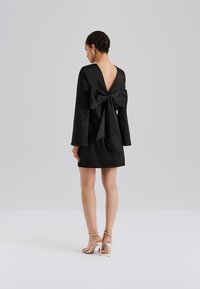 Svart kjole med lange ermer, dyp V-utringning bak, og stor sløyfedetalj. Glatt stoff, tettsittende fasong, midtlengde til låret. Par med sølvfargede stroppesandaler.