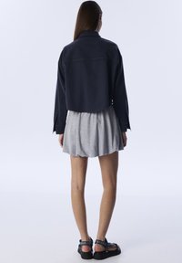 Gekürzte marineblaue Denimjacke mit Kragen und Nähten, kombiniert mit einem grauen Faltenrock. Schwarze Sandalen mit Verzierungen.