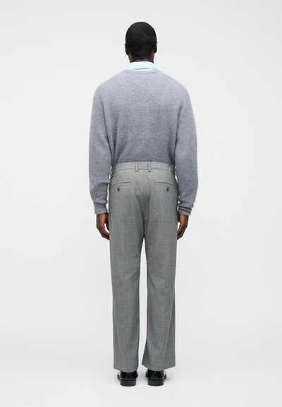 Άνδρας που κοιτάζει προς τα πίσω, φοράει ανοιχτό γαλάζιο πουκάμισο με γιακά, γκρι χνουδωτό πουλόβερ, γκρι καλοραμμένο παντελόνι και μαύρα επίσημα παπούτσια, μπροστά σε απλό φωτεινό φόντο.