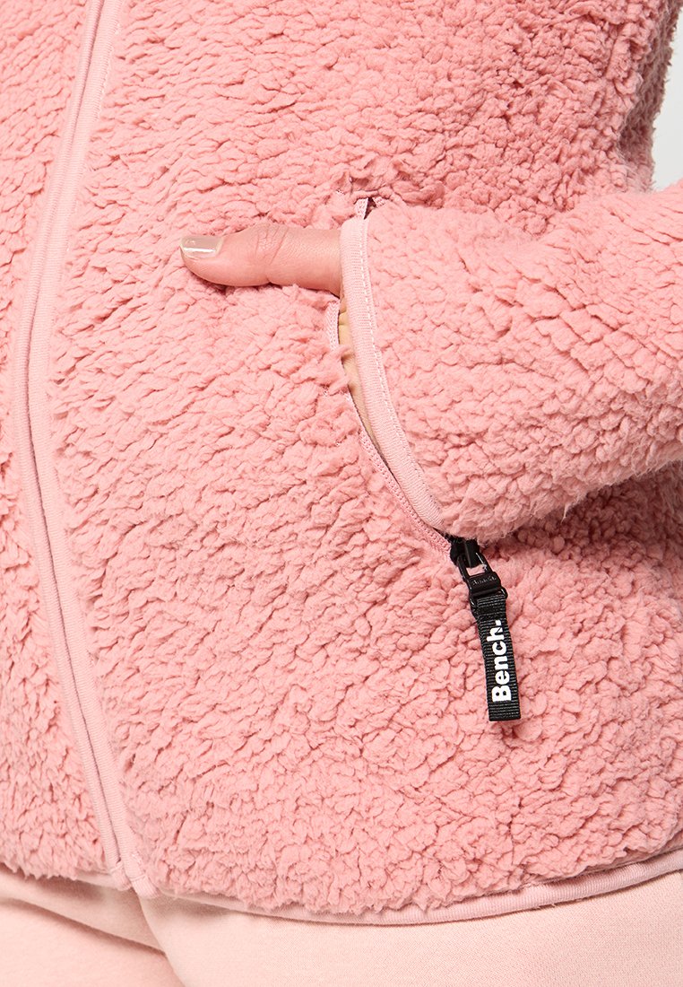 Różowa polarowa kurtka o teksturowanej, puszystej powierzchni; posiada zamek błyskawiczny z przodu oraz boczne kieszenie; markowy uchwyt zamka.