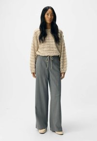 Beiger Strickpullover mit strukturiertem Muster, kombiniert mit hellgrauen Streifen-Hosen mit Kordelzugbund. Braune Slipper.
